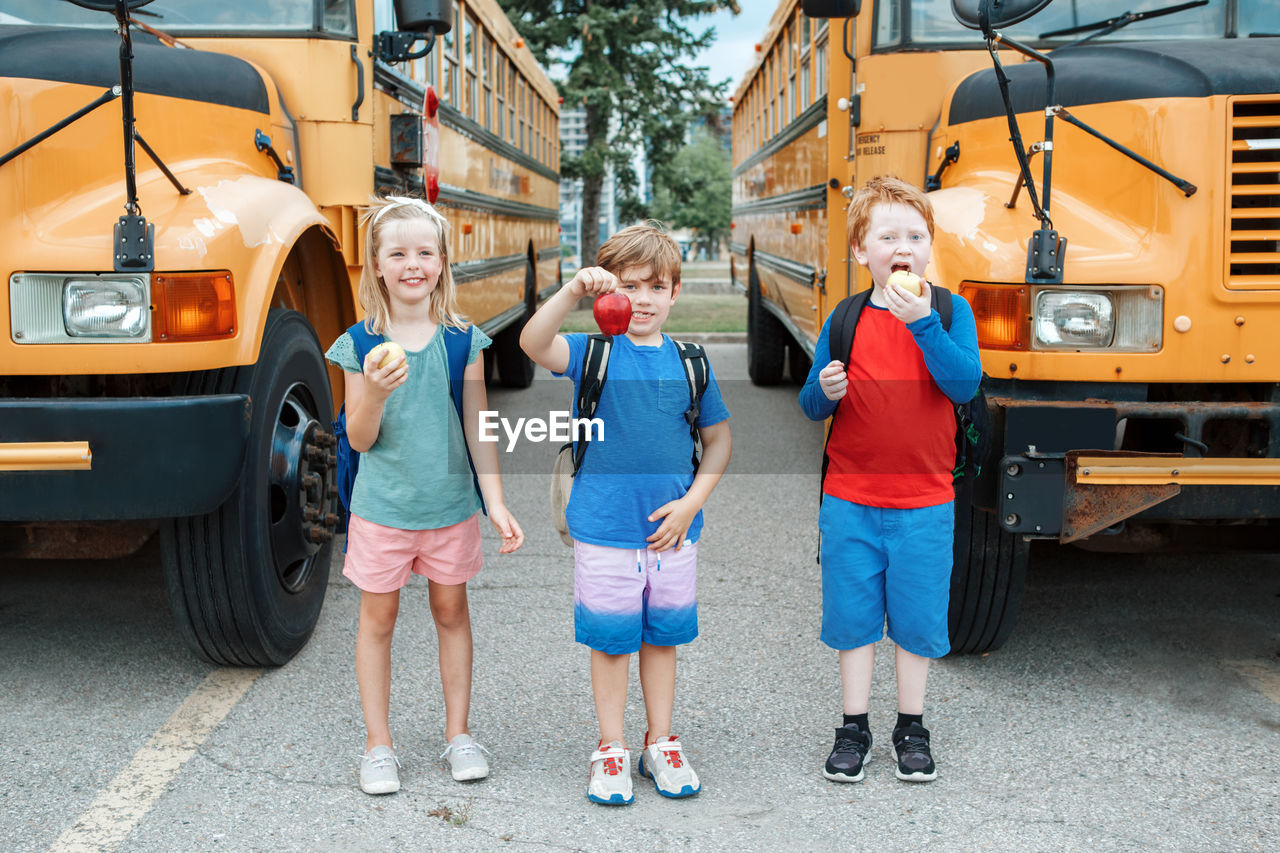 Children boys and girl students friends eating apples healthy snack by yellow school bus outdoors. 