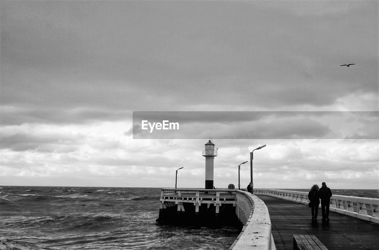 PEOPLE STANDING BY LIGHTHOUSE BY SEA AGAINST SKY