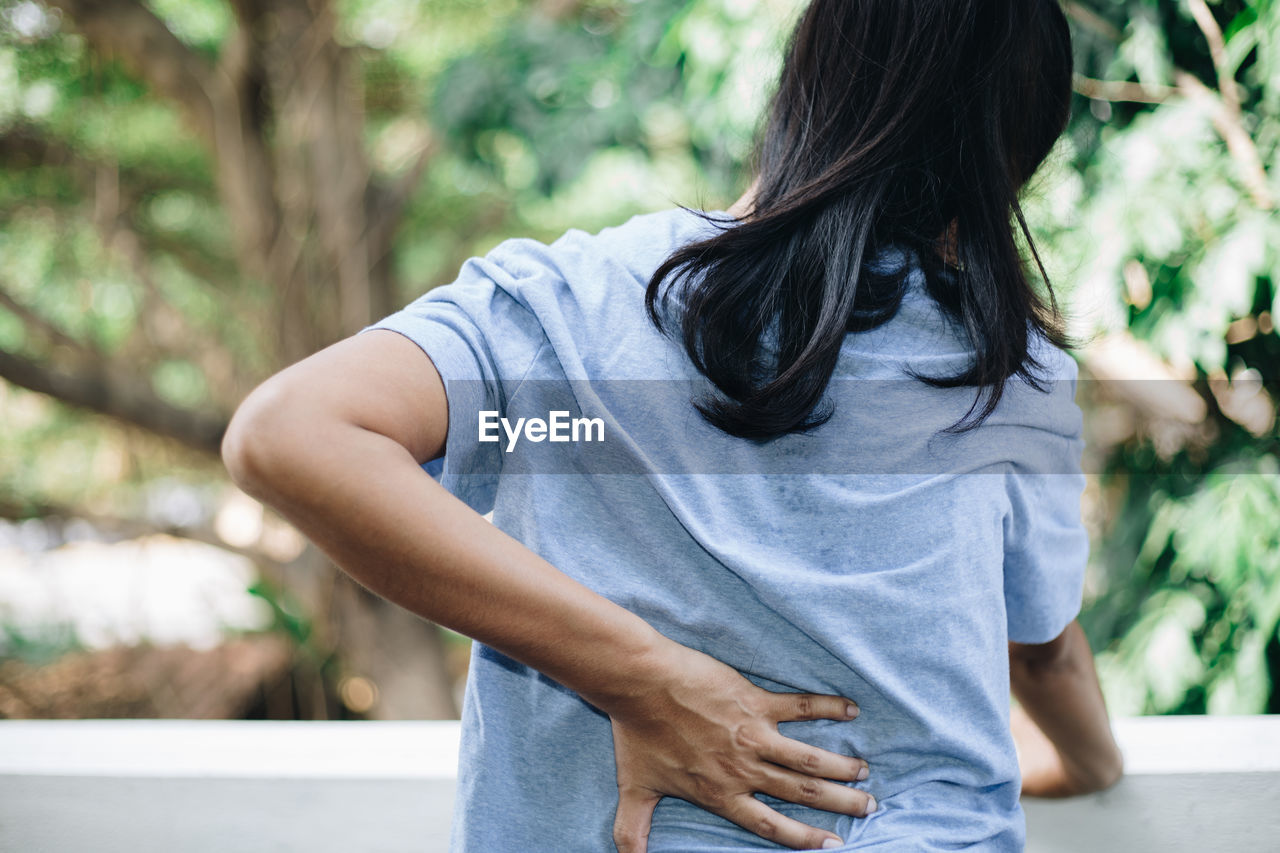 Rear view of woman touching back in pain while standing against tree