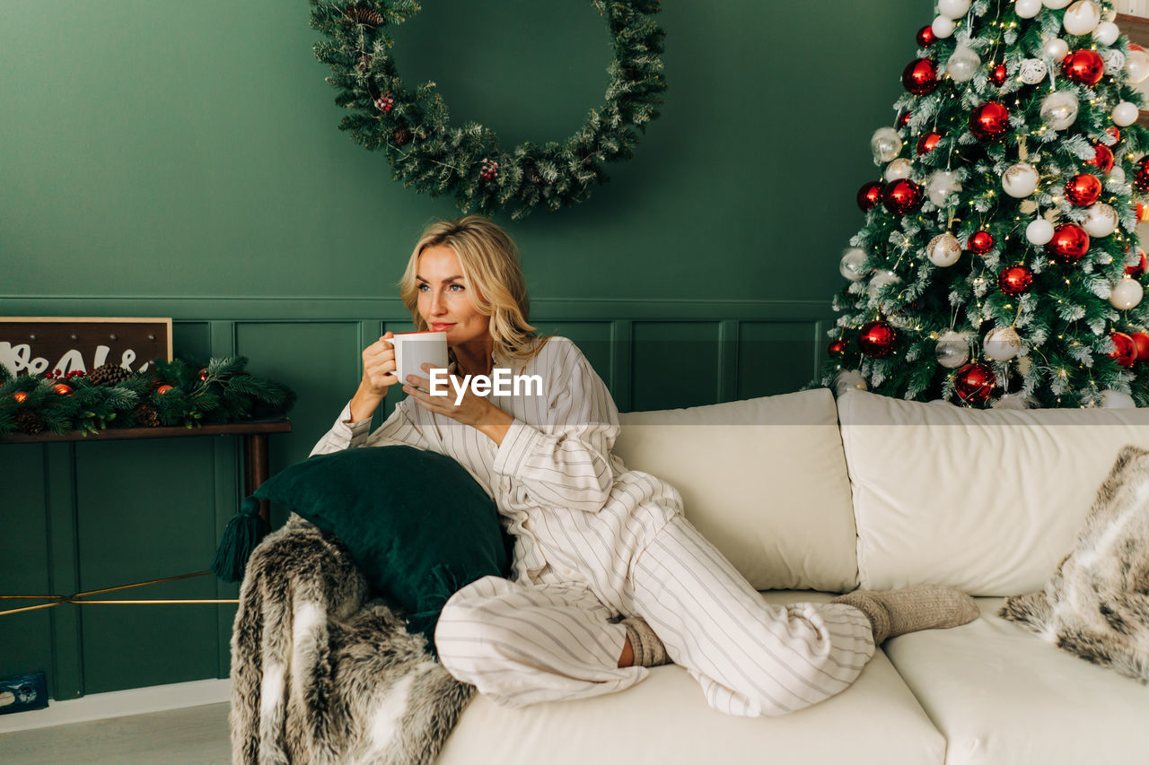 Gorgeous caucasian blond woman sitting on sofa on christmas morning drinking coffee enjoying