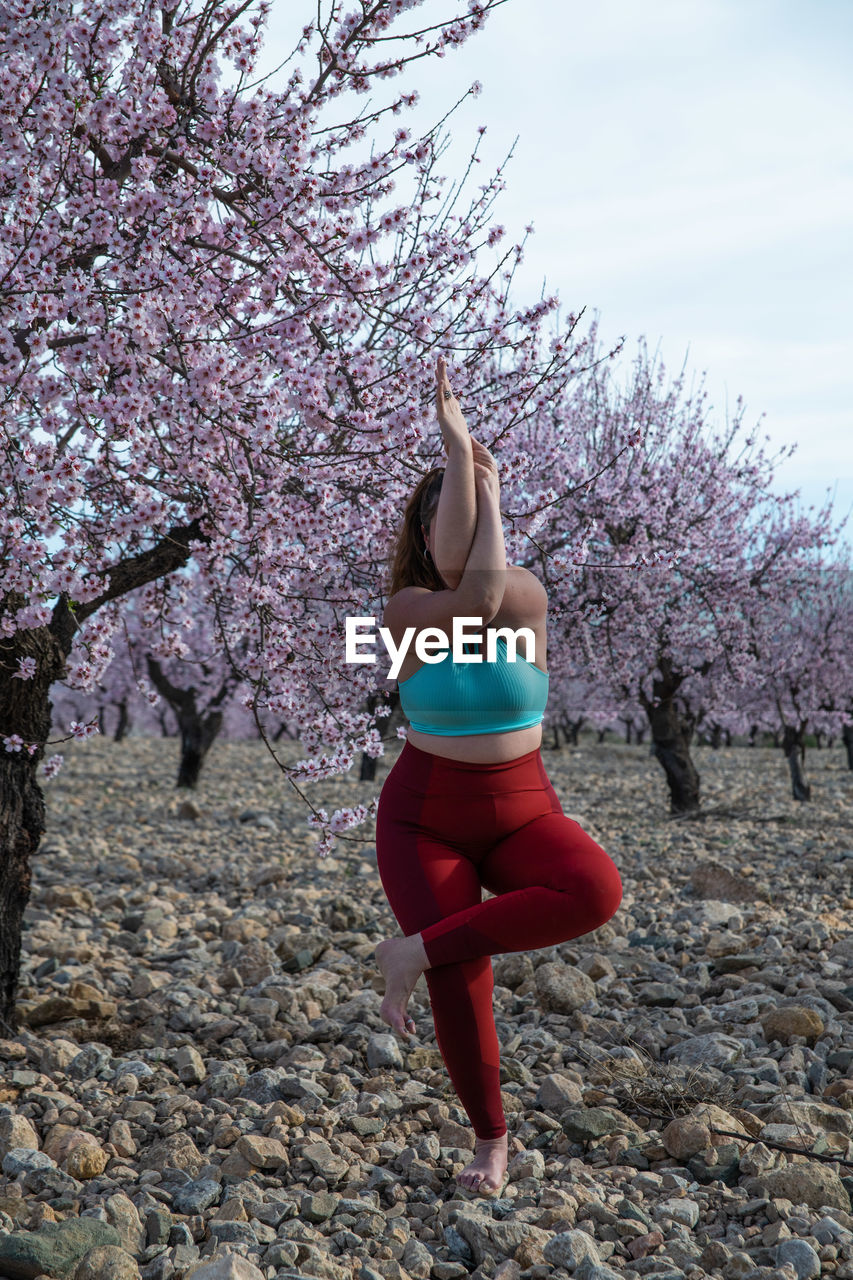 Unrecognizable curvy female in sportswear standing in garudasana in garden with blossoming almond trees and practicing yoga while balancing on leg