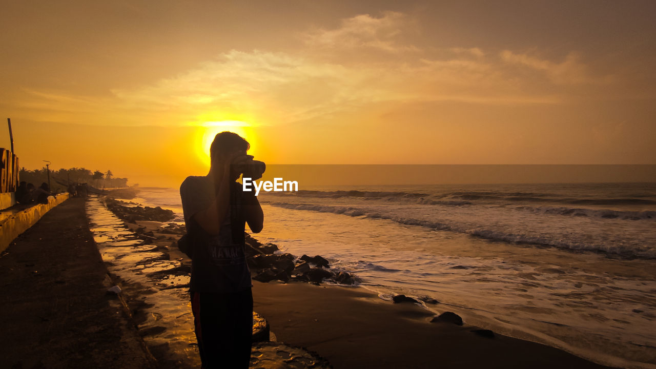 Man photographing while standing on beach against sky during sunset