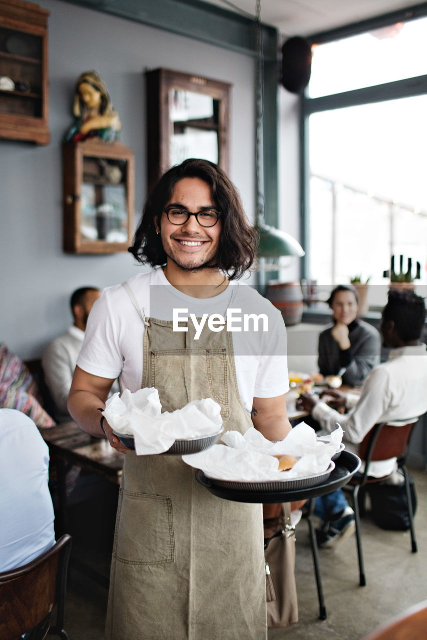 Portrait of confident young waiter serving food while standing against customers at restaurant