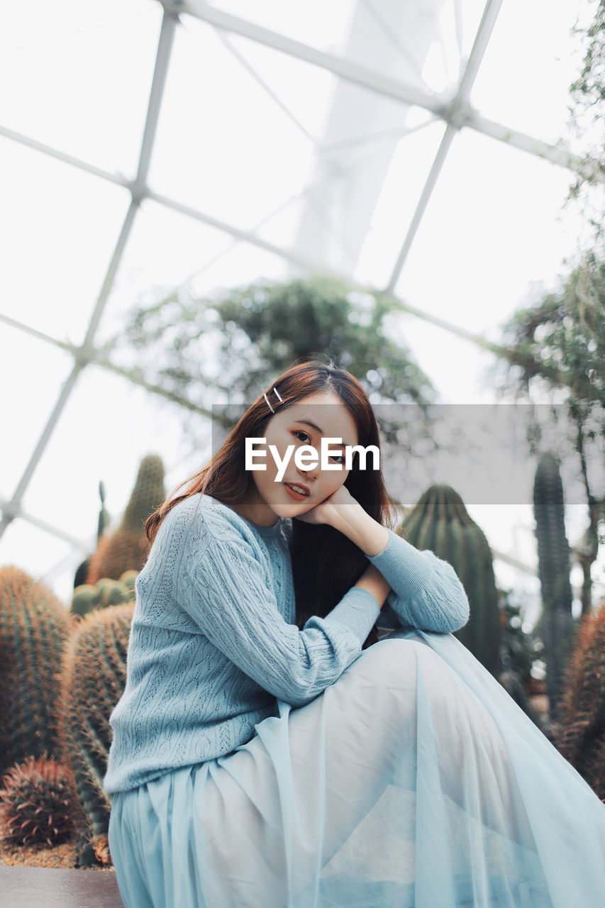 Portrait of young woman sitting against plants in greenhouse