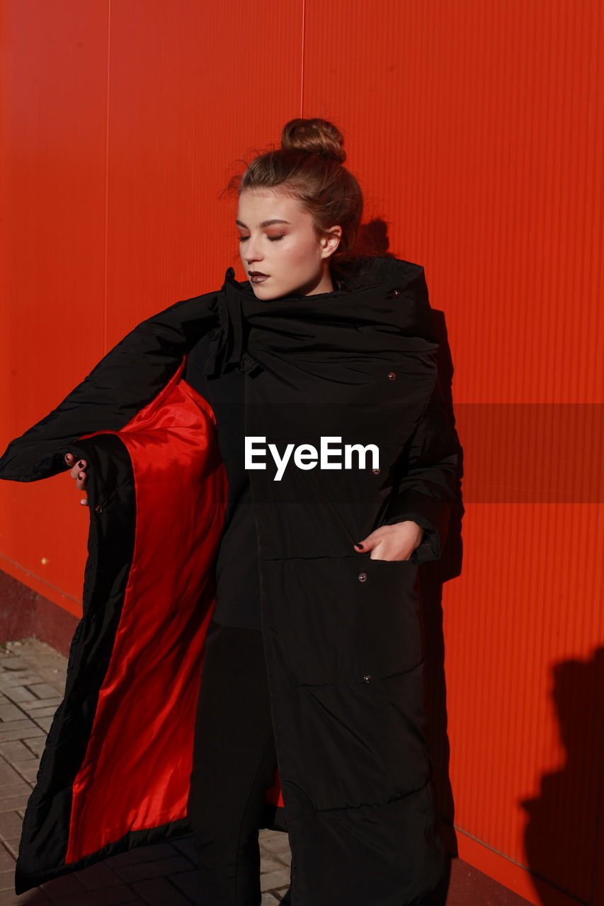 Fashionable teenage girl wearing black warm clothing standing against red wall during sunny day