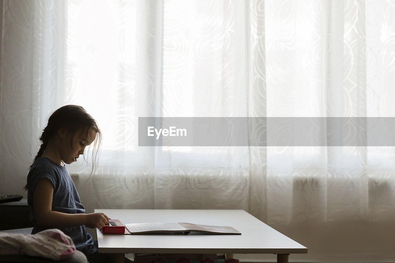 Side view of girl playing toy piano on table by window at home