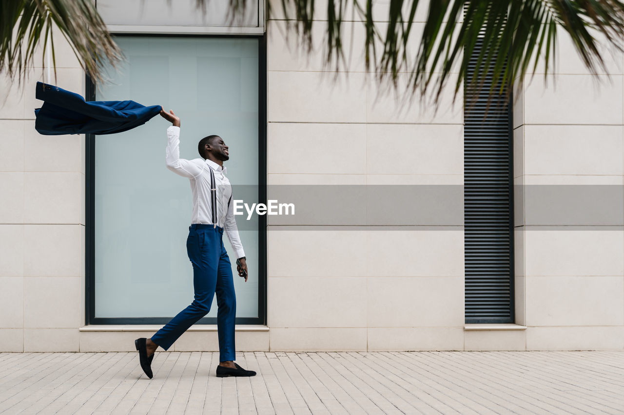 Happy male entrepreneur holding blue suit while walking on sidewalk in city