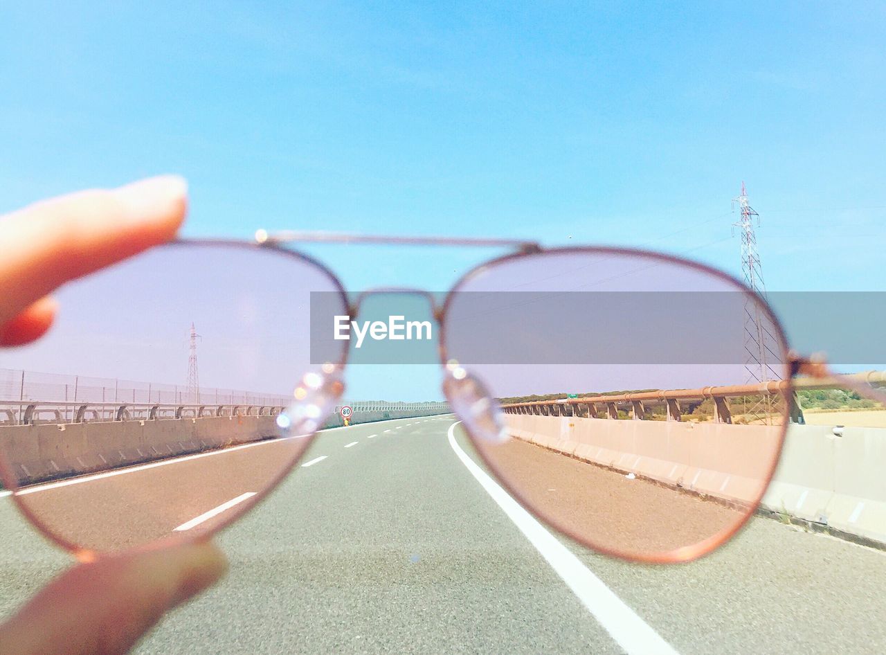 Cropped hand holding sunglasses over road against clear sky