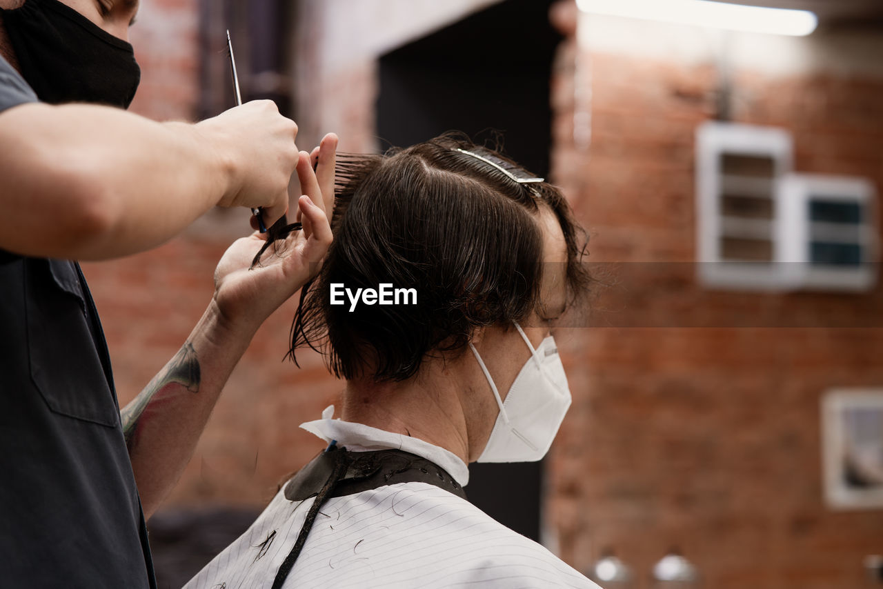 Barber in protective mask and latex glove styling adult man in a barbershop salon. new normal