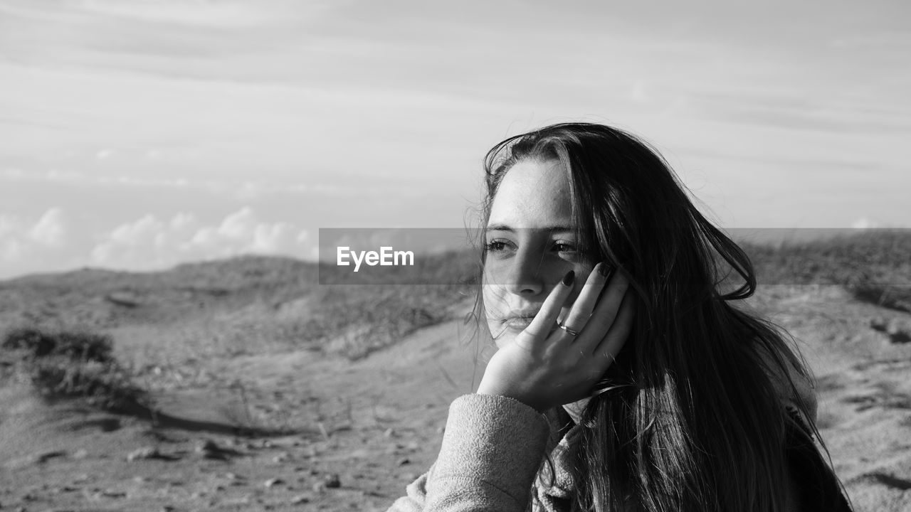 Thoughtful beautiful woman with hand on chin sitting against sky