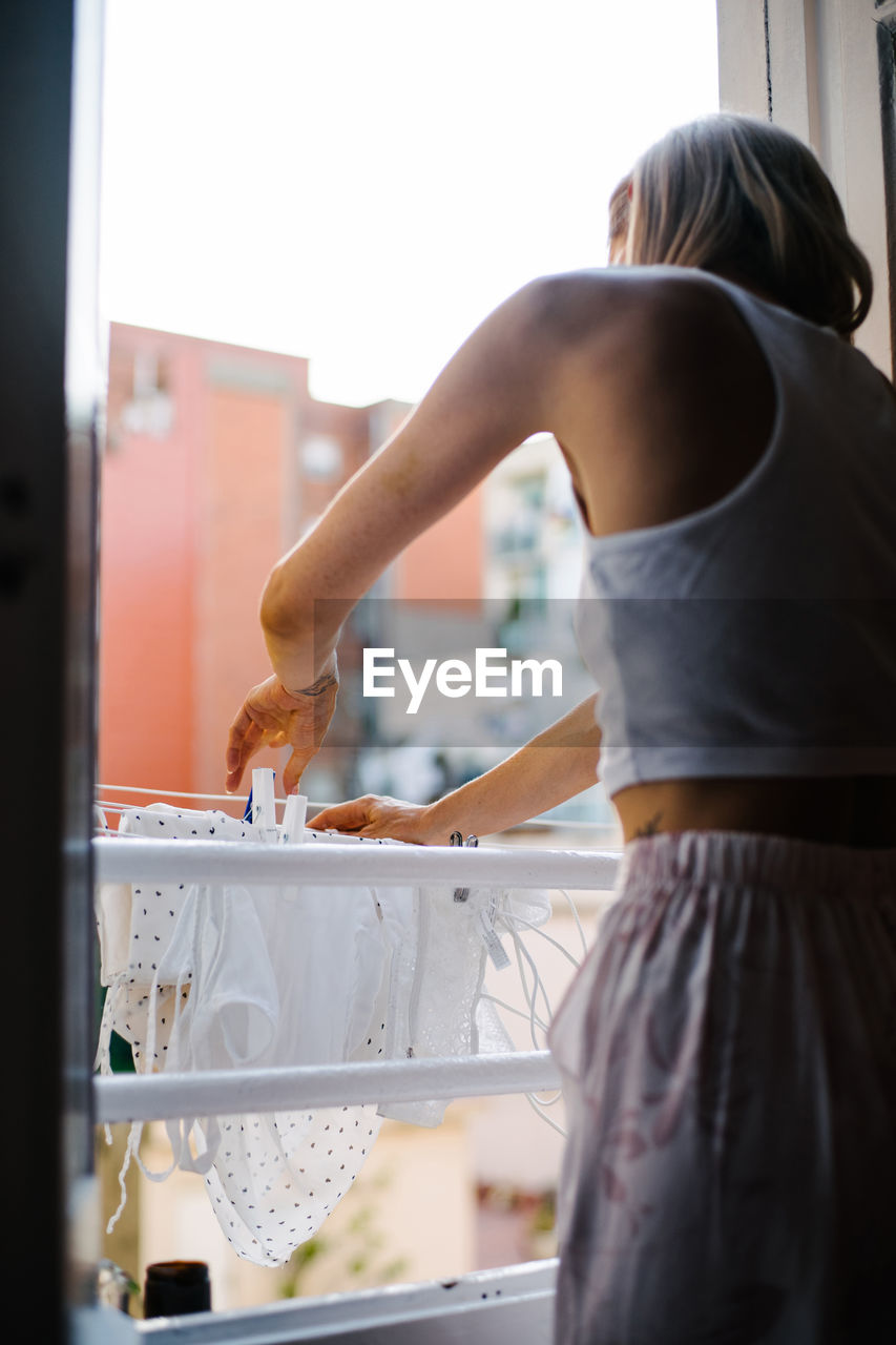 Back view of unrecognizable female in domestic outfit standing near window and attaching wet clothes with clothespins while doing household chores at weekend