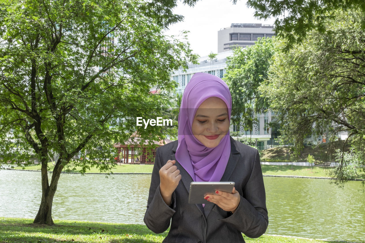 Young woman with hijab sitting and texting her friends using her tablet at park during sunny day