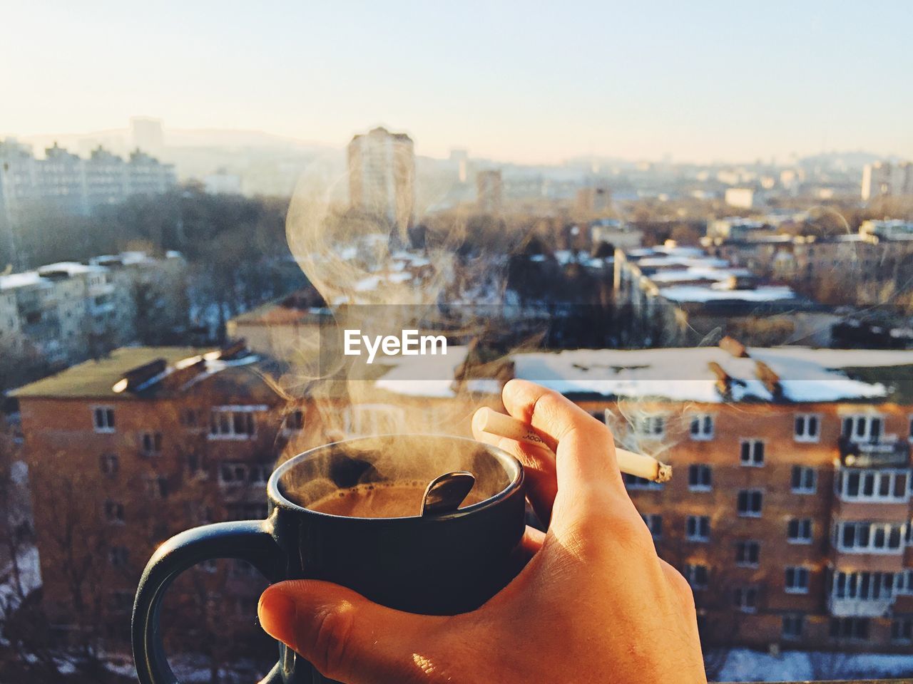 Cropped hand holding coffee cup and cigarette against cityscape