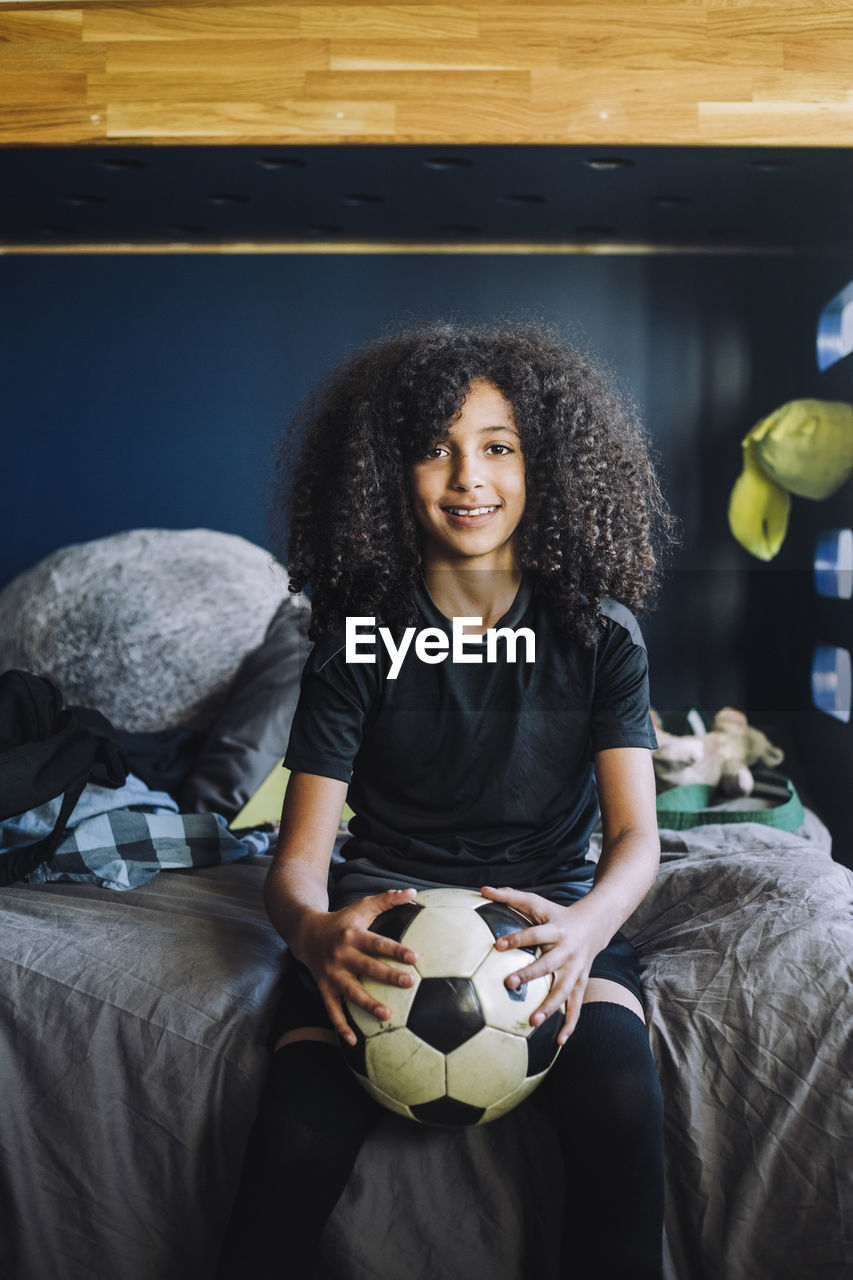 Portrait of curly hair girl with soccer ball sitting on bed at home