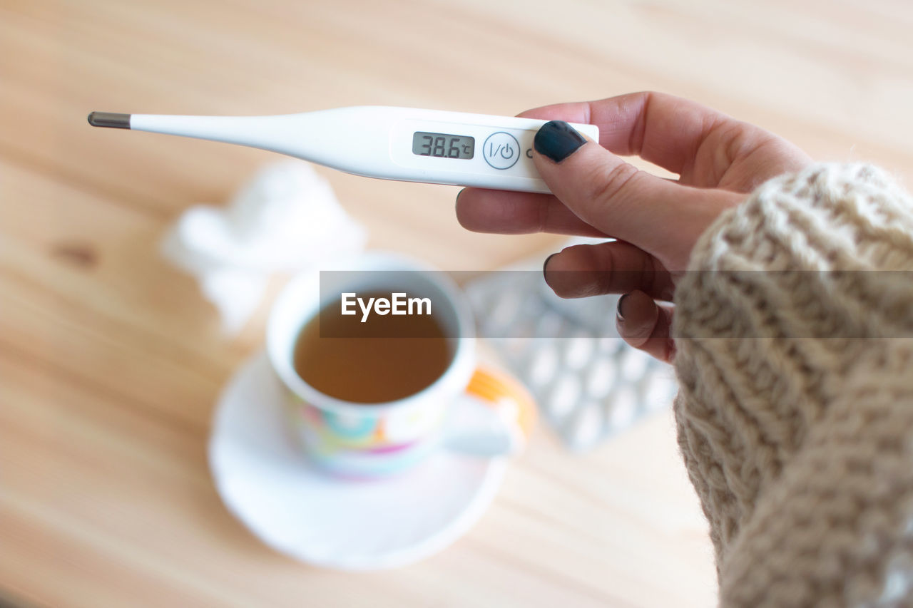 Cropped hand of woman holding thermometer against coffee cup on table