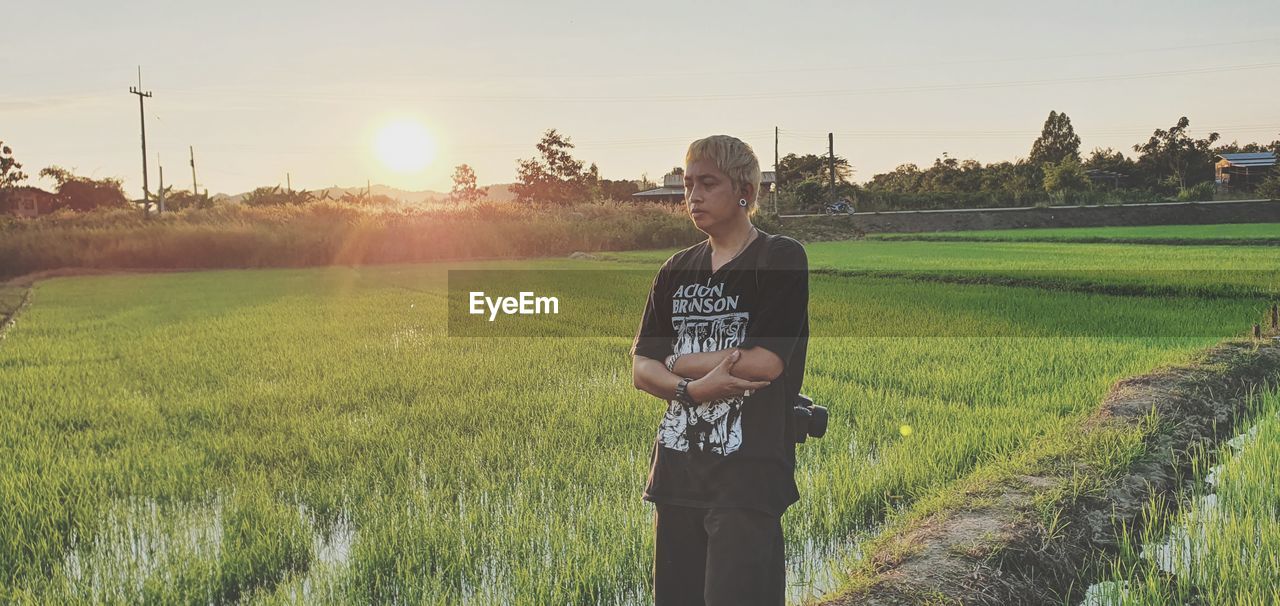 agriculture, field, plant, sky, nature, one person, adult, landscape, sunlight, rural area, land, crop, lens flare, rural scene, grass, sunset, growth, standing, sun, environment, tree, men, farm, meadow, prairie, lifestyles, three quarter length, back lit, outdoors, person, paddy field, beauty in nature, sunbeam, day, clothing, occupation, sports, grassland, green, leisure activity