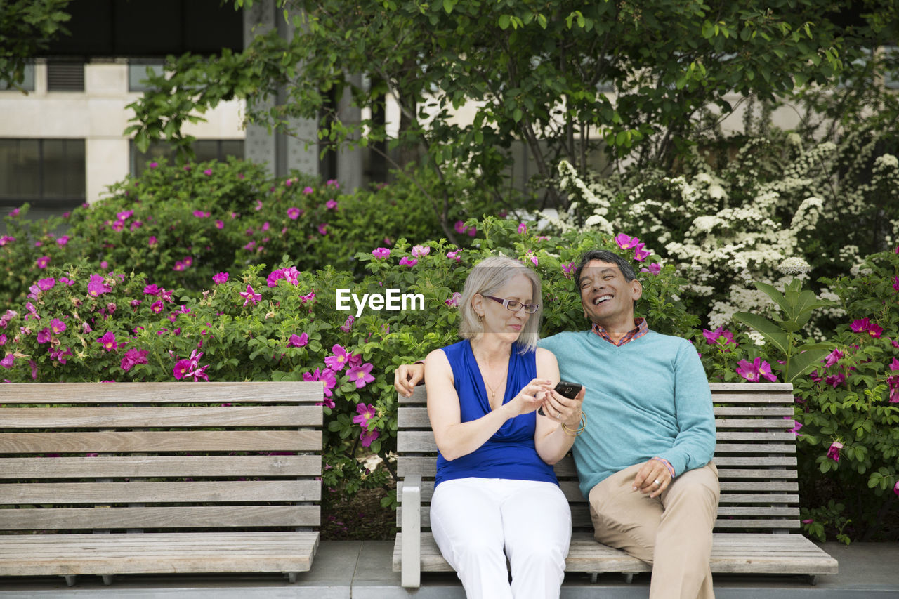 Happy mature couple using smart phone sitting on bench in park