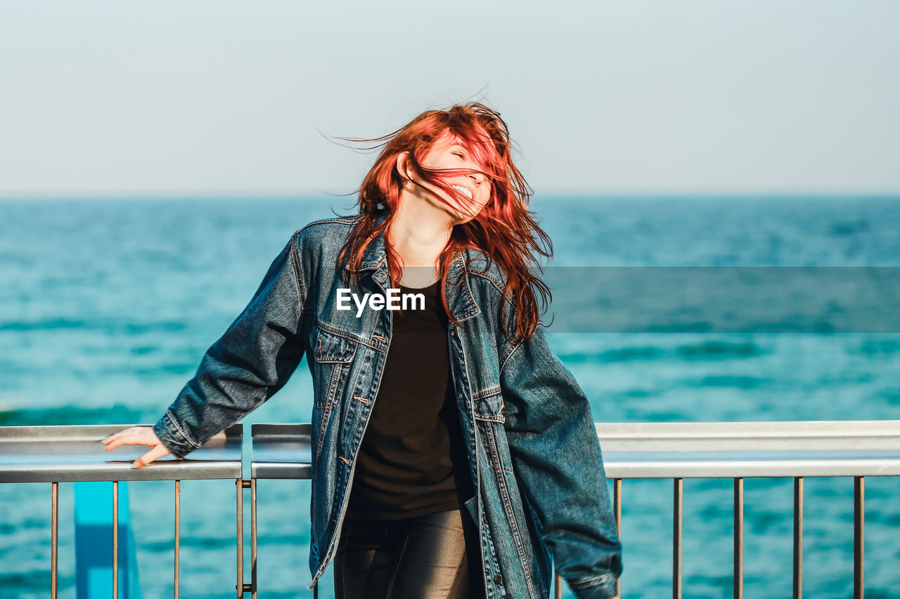 Smiling young woman with tousled hair standing by railing against sea