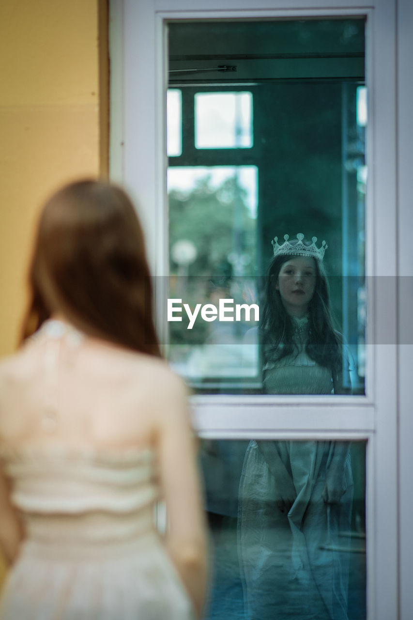 A girl with a crown on her head looking at her reflection in the window
