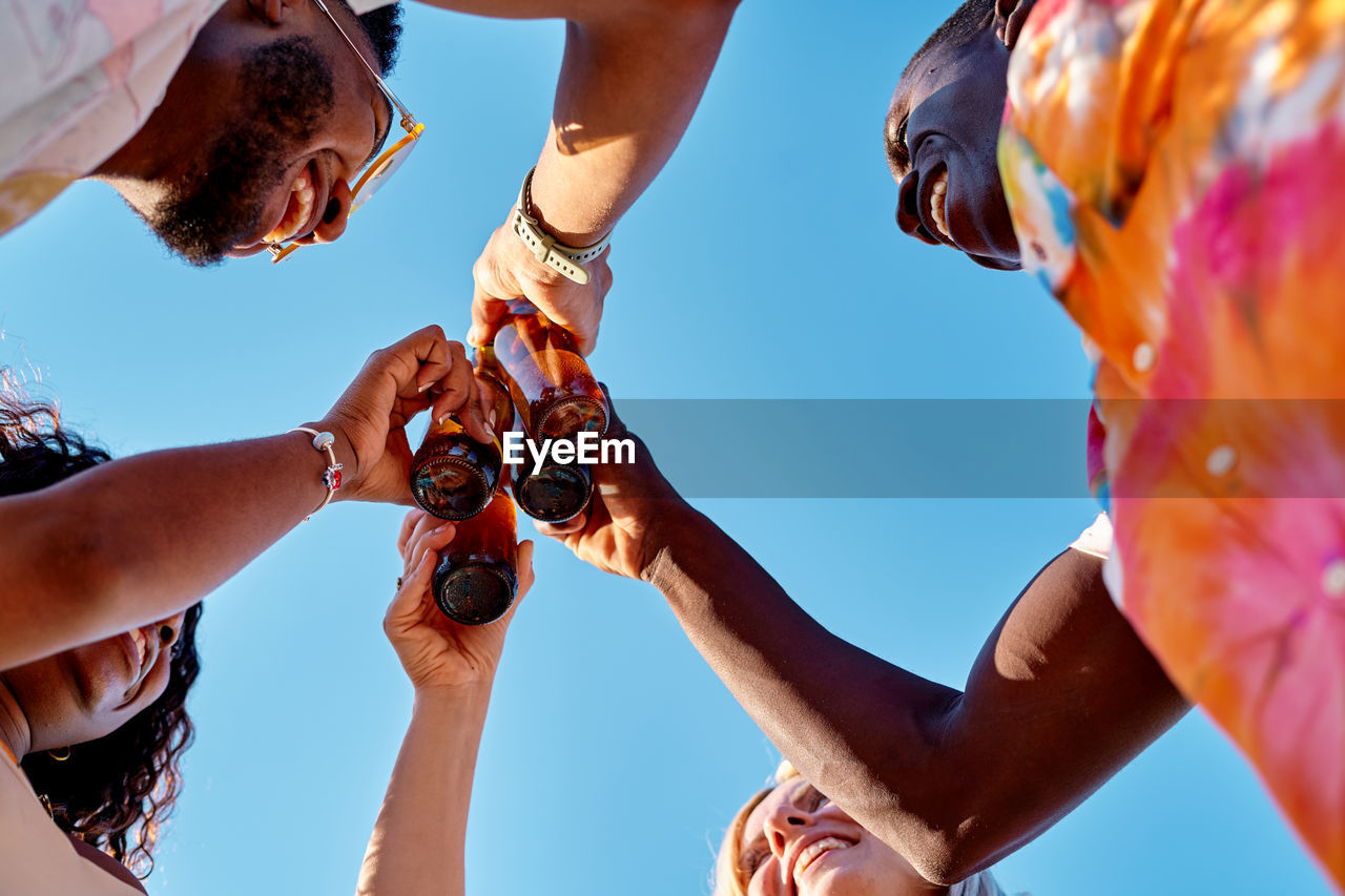 From below of cheerful young diverse best friends smiling and toasting with bottles of beer against cloudless blue sky on sunny summer day