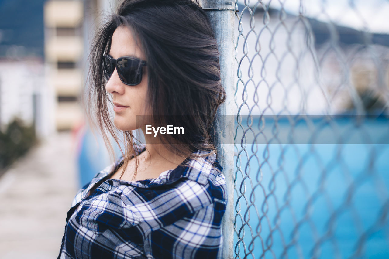 Beautiful young woman wearing sunglasses leaning on chainlink fence