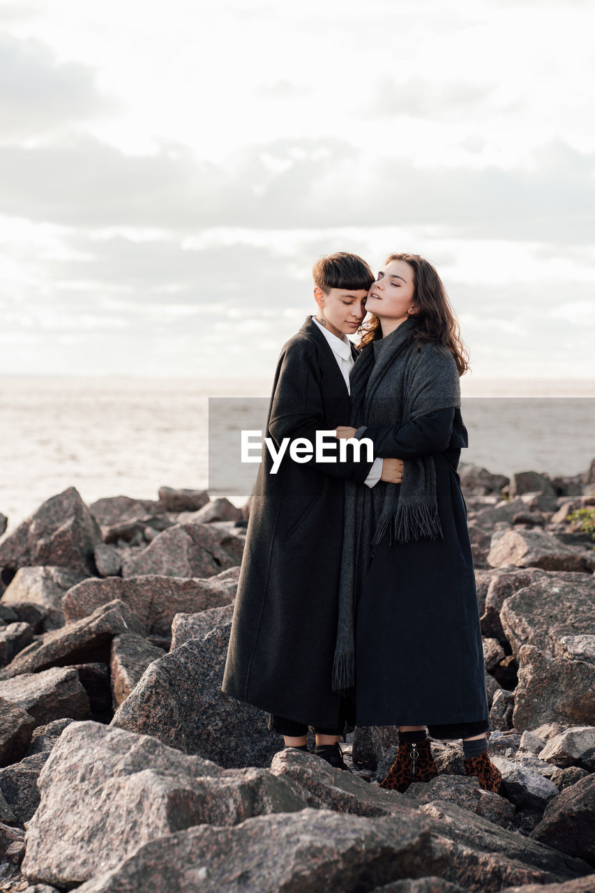 Lesbian women embracing while standing on rock against sea and sky