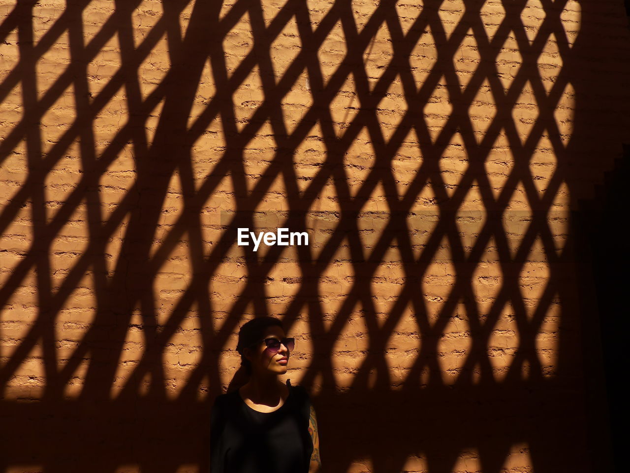 Woman wearing sunglasses looking away standing against shadow pattern on wall