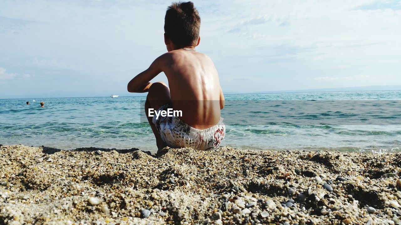 Shirtless boy sitting at beach against sky