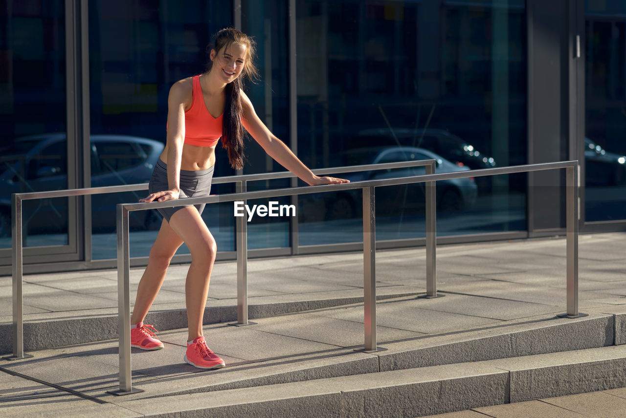 Full length of young woman doing exercise by railing on sunny day