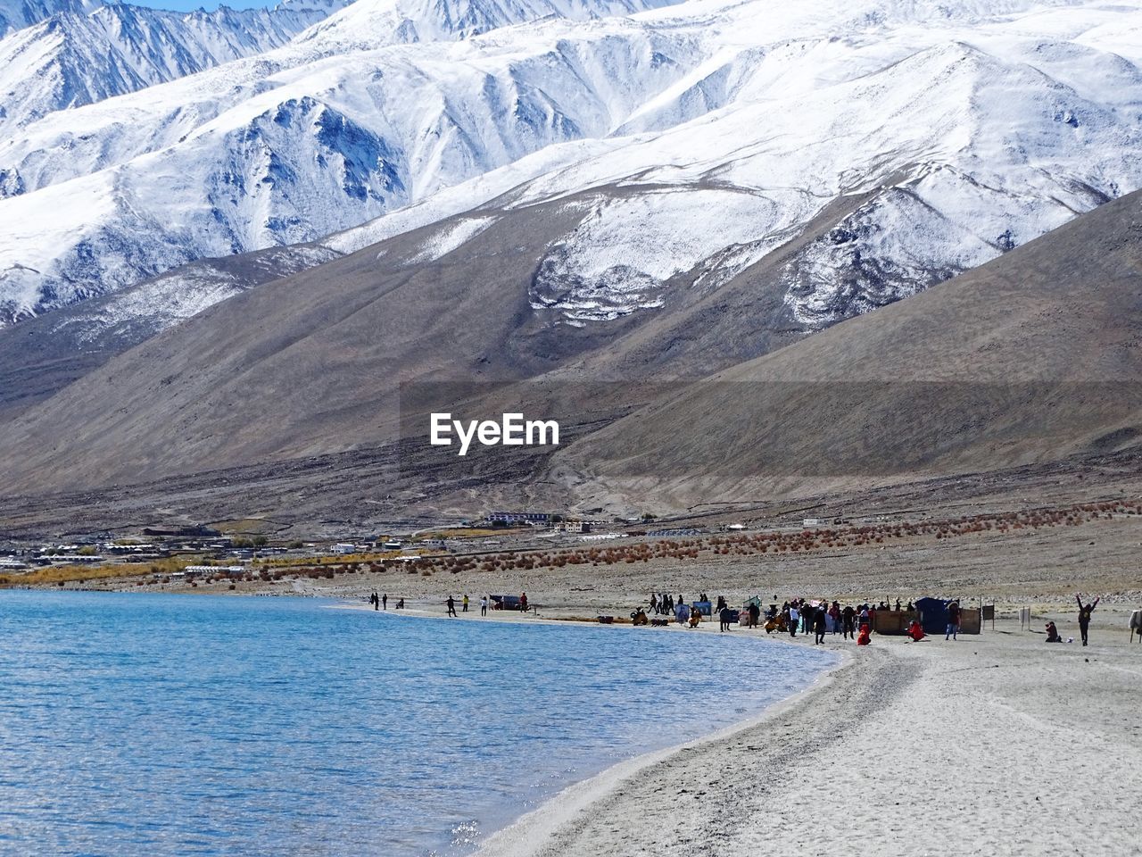 Group of people on lakeshore against snowcapped mountain