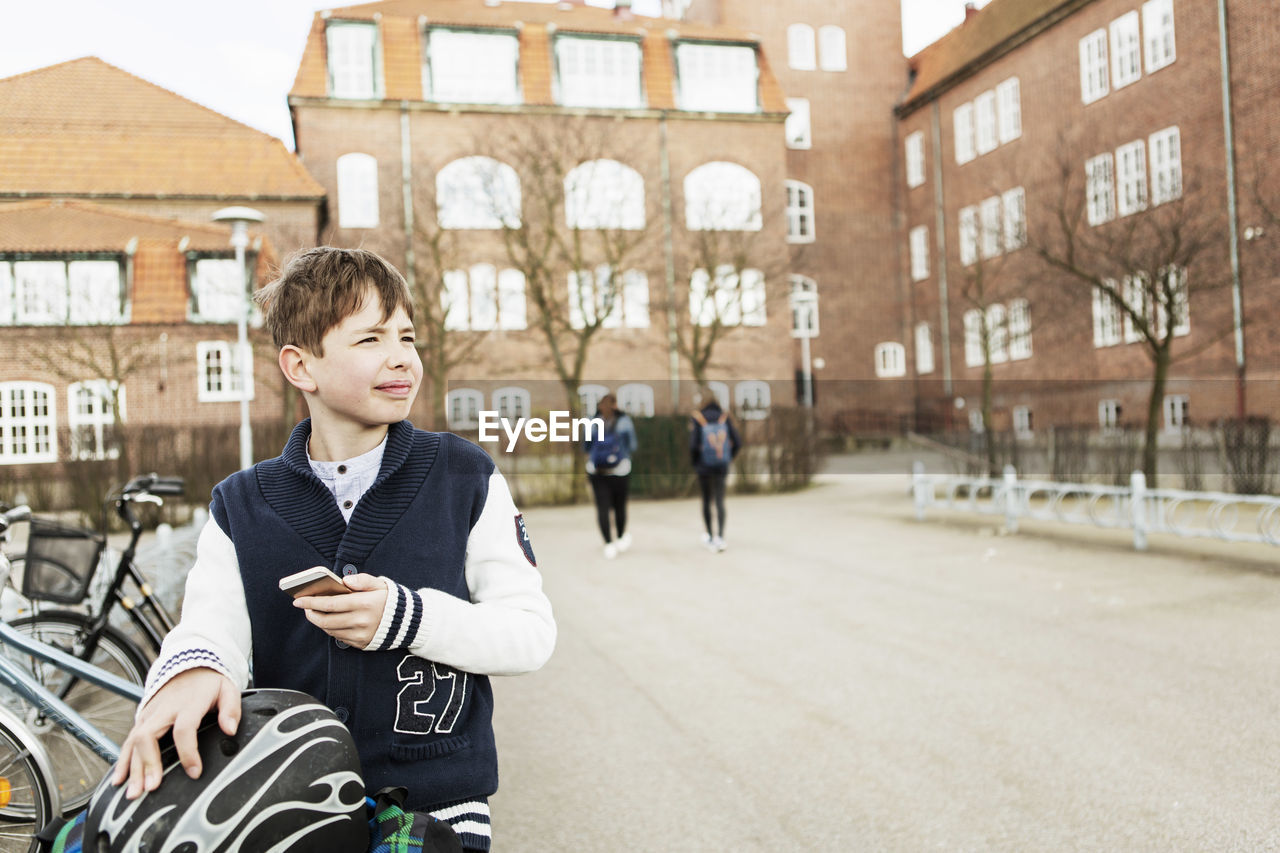 Boy looking away while holding mobile phone outside school building