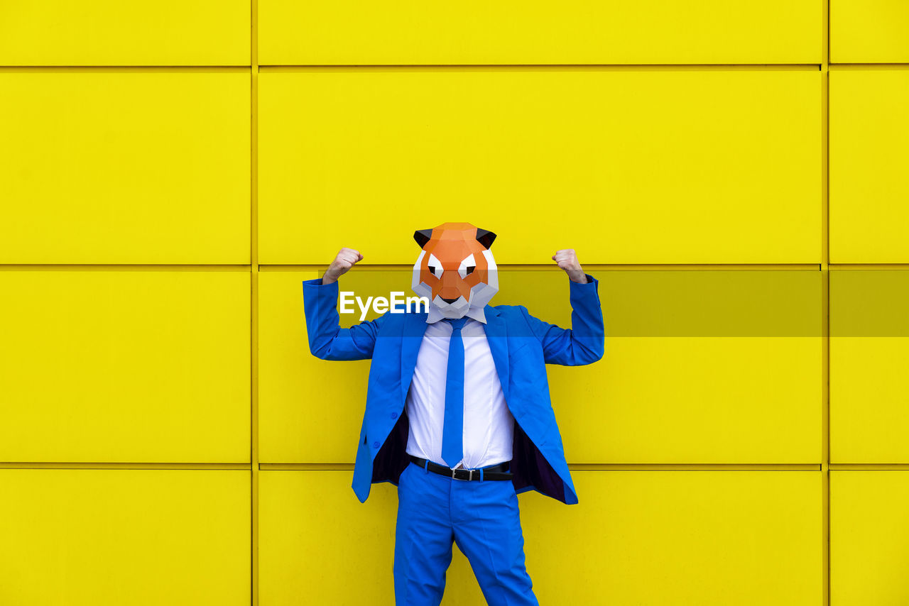 Man wearing vibrant blue suit and tiger mask flexing in front of yellow wall