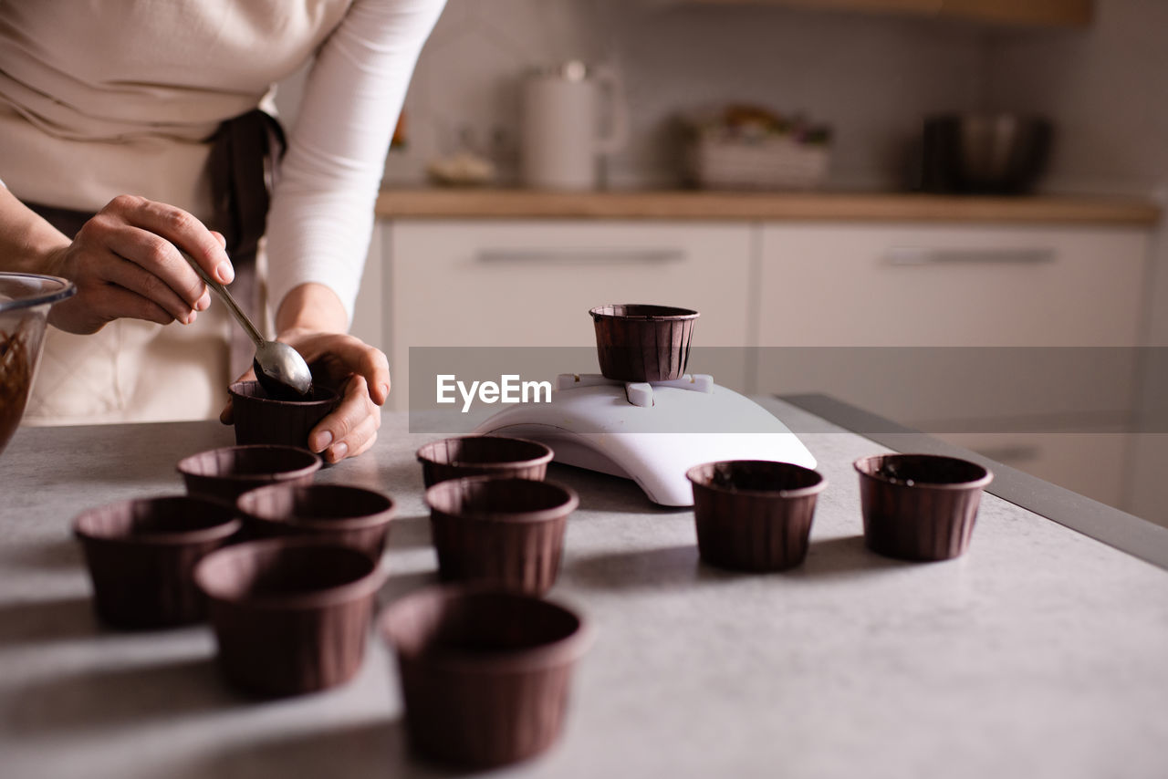 Woman making chocolate cupcakes weigh forms on kitchen scale on table close up.