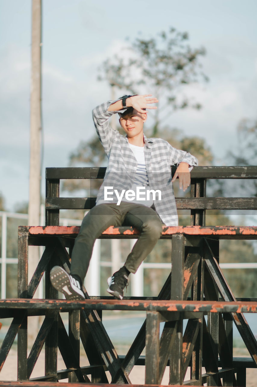 FULL LENGTH OF YOUNG MAN SITTING ON RAILING AGAINST BLURRED BACKGROUND