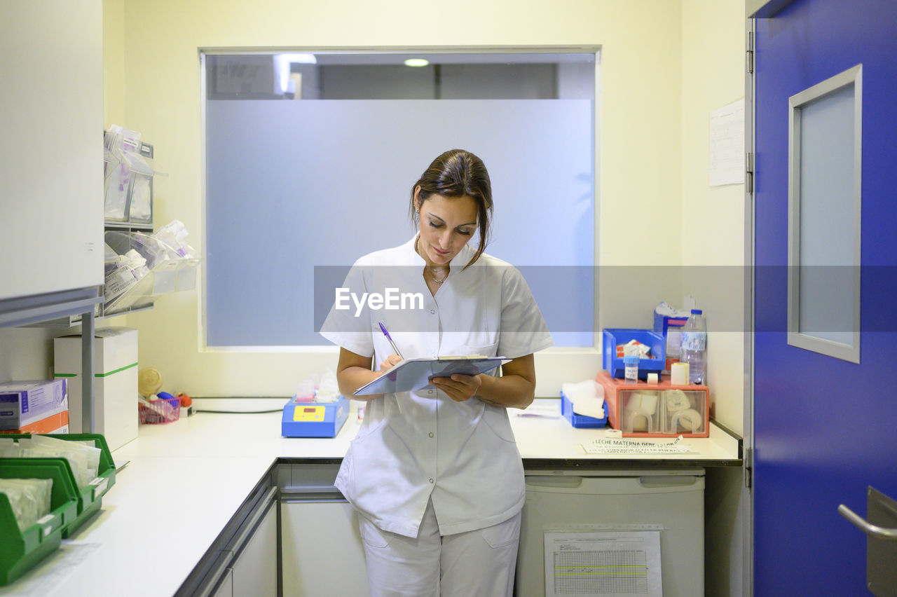 Woman in medical uniform writing on clipboard while standing in office during shift in children hospital