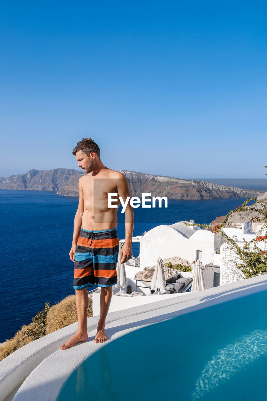 Shirtless man standing by swimming pool over sea against clear blue sky