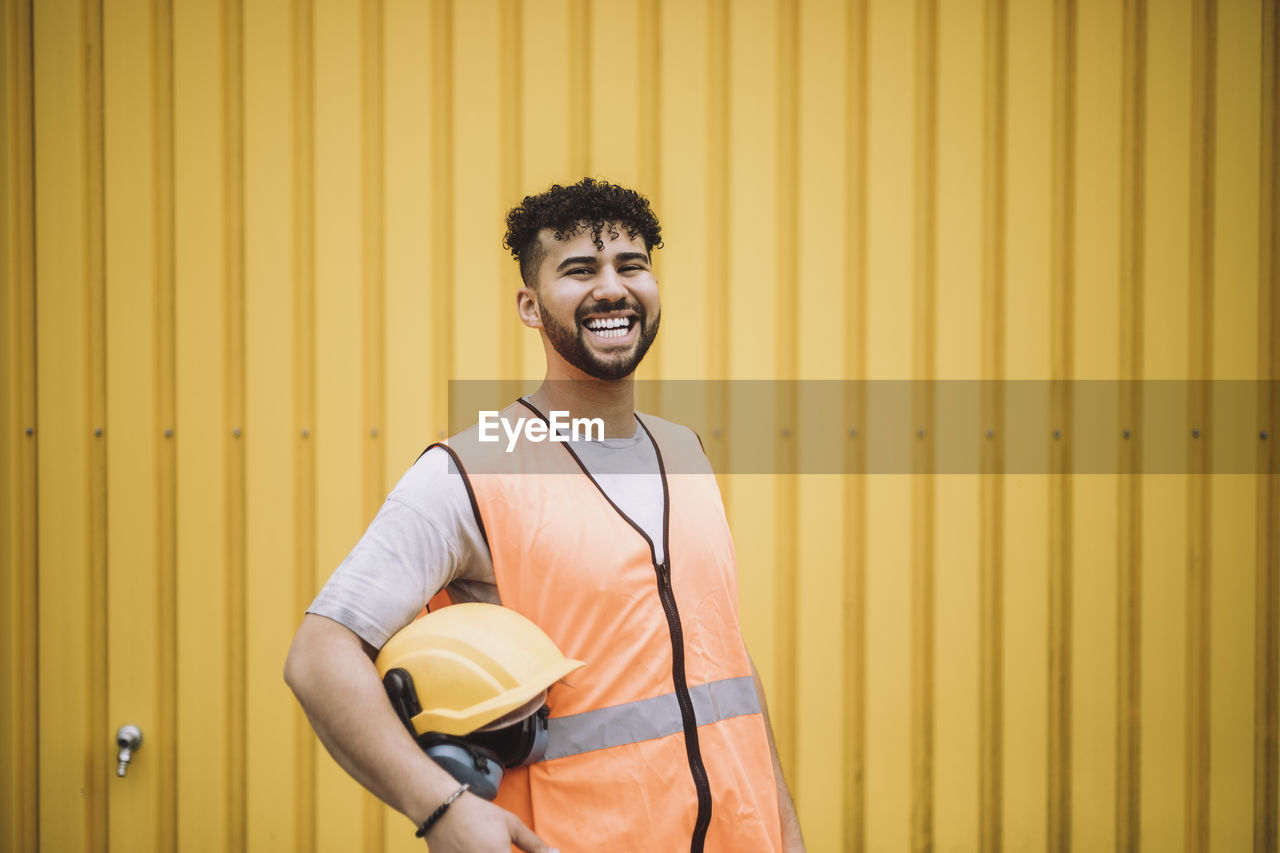 Portrait of cheerful young construction worker with hardhat standing against yellow metal wall