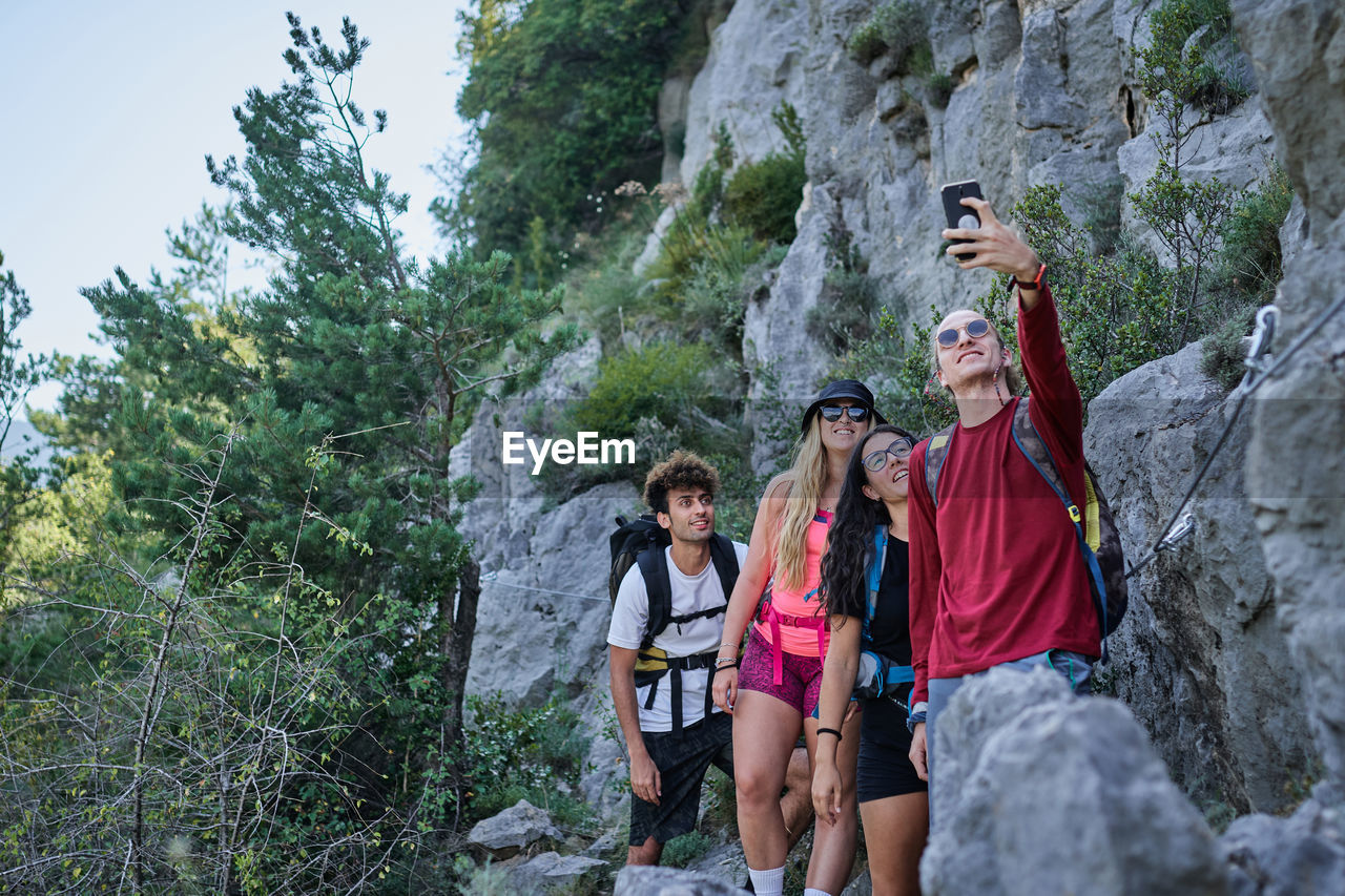 Group of friendly tourists with backpacks standing in highland area and taking photo on selfie camera during vacation