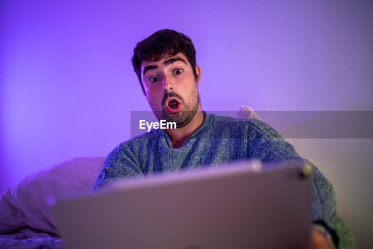 Man using digital tablet sitting on bed against wall at home