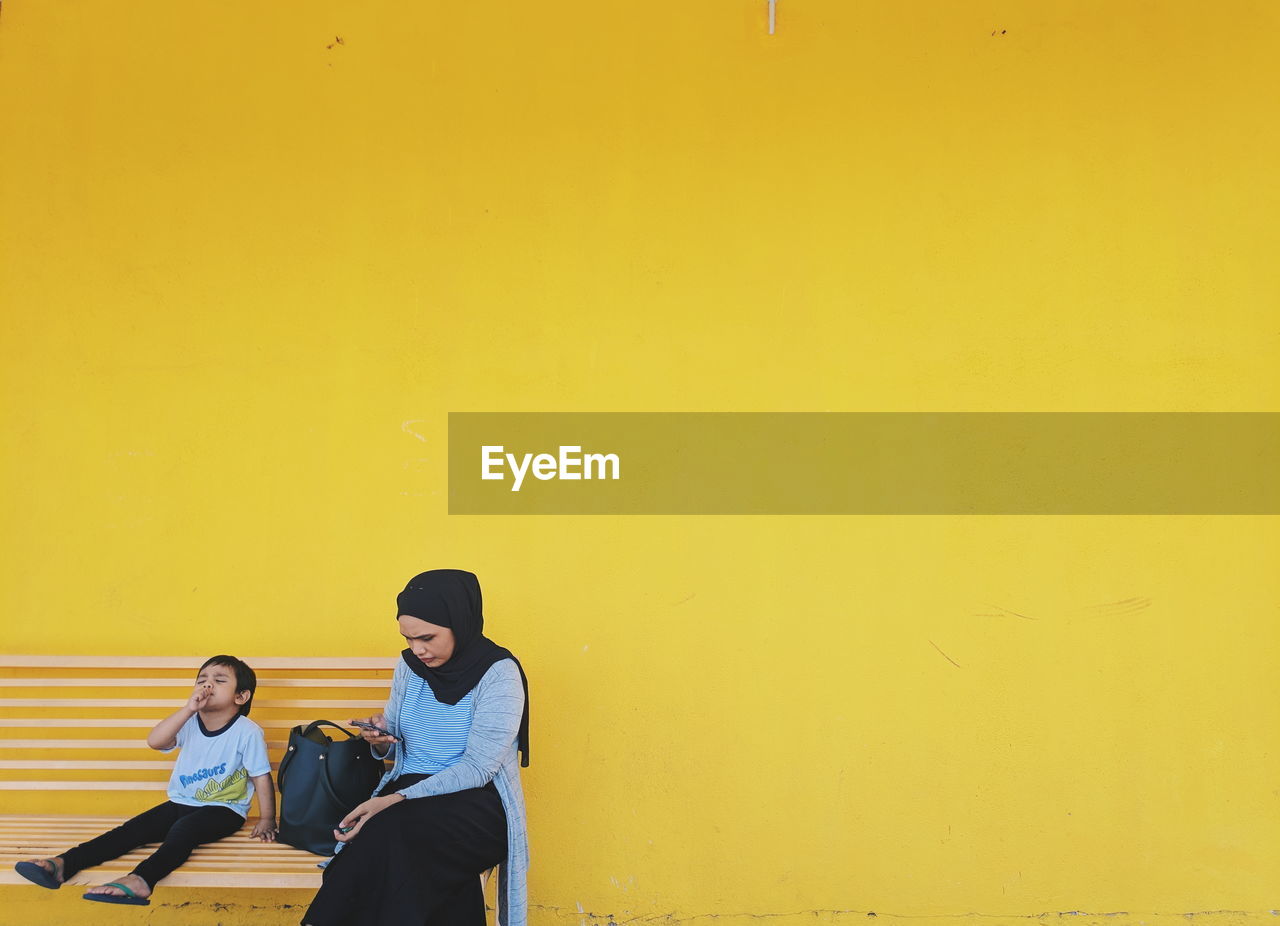 Mother and son sitting on bench against yellow wall