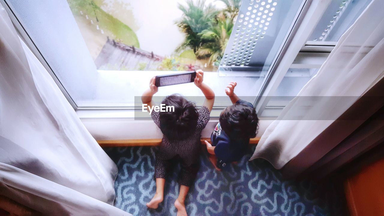 High angle view of siblings photographing by window at home