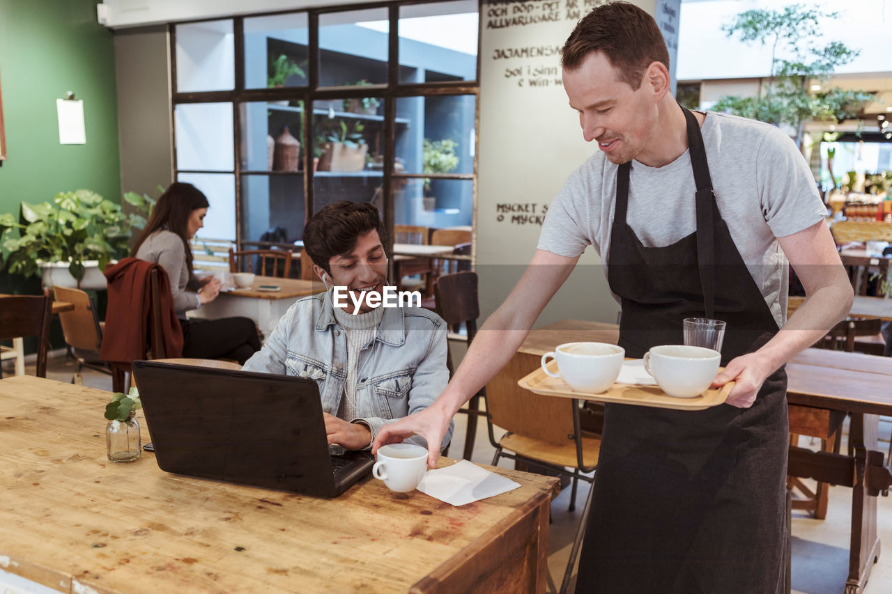 Mid adult owner serving coffee to male customer at cafe