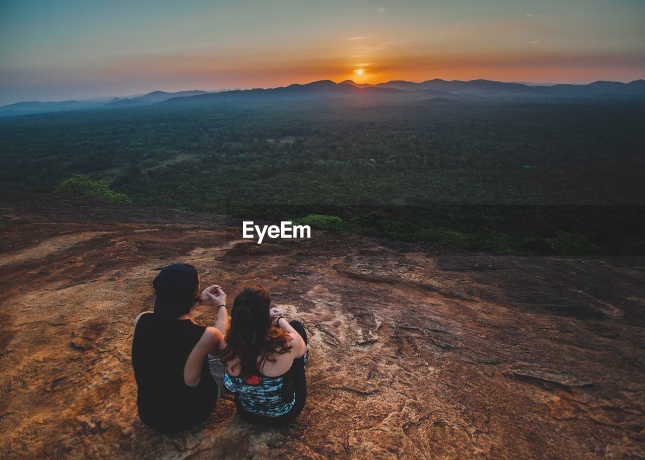 PEOPLE SITTING ON LANDSCAPE AGAINST SKY DURING SUNSET