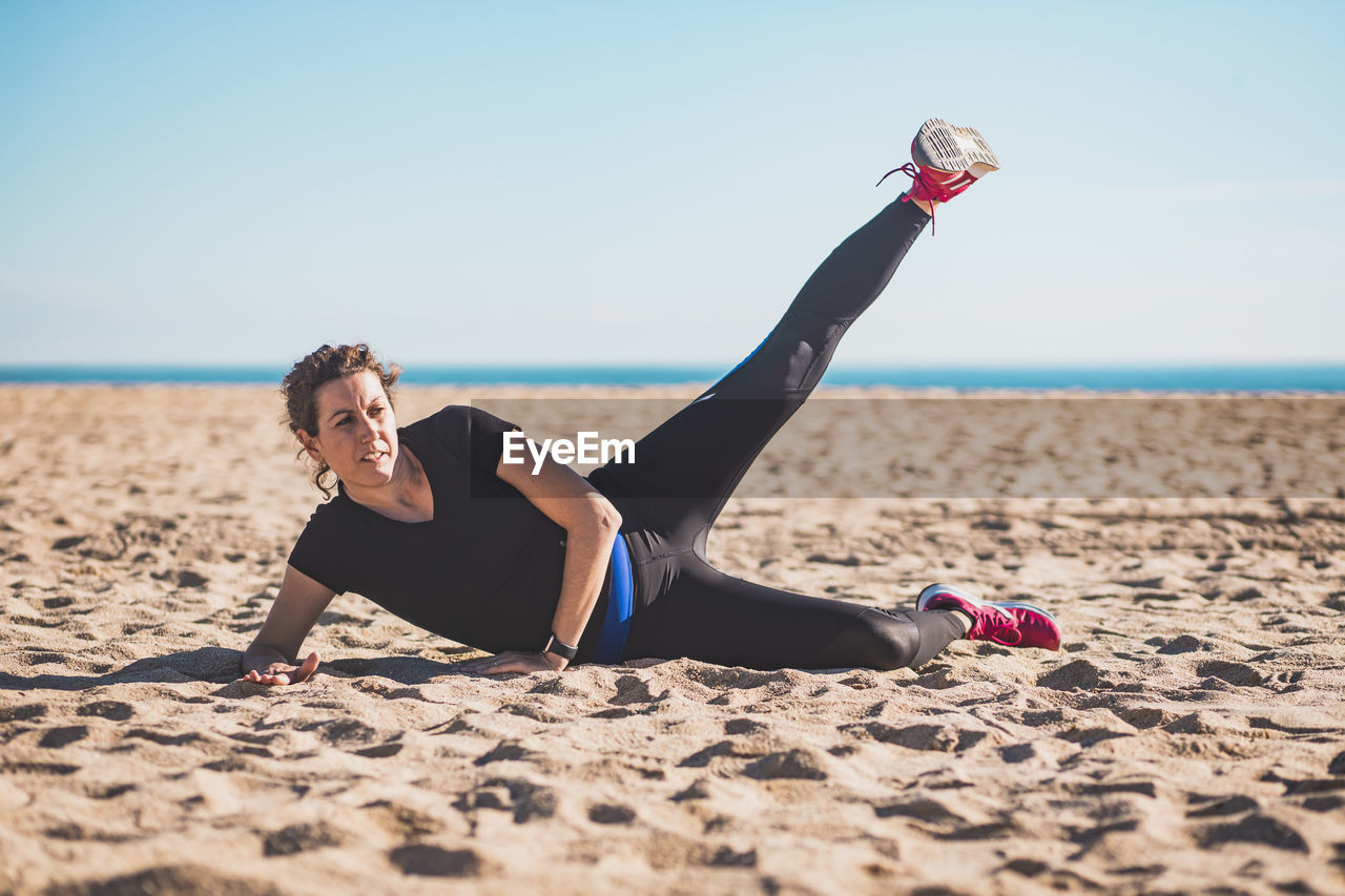 Full length of woman exercising while lying on beach against sky