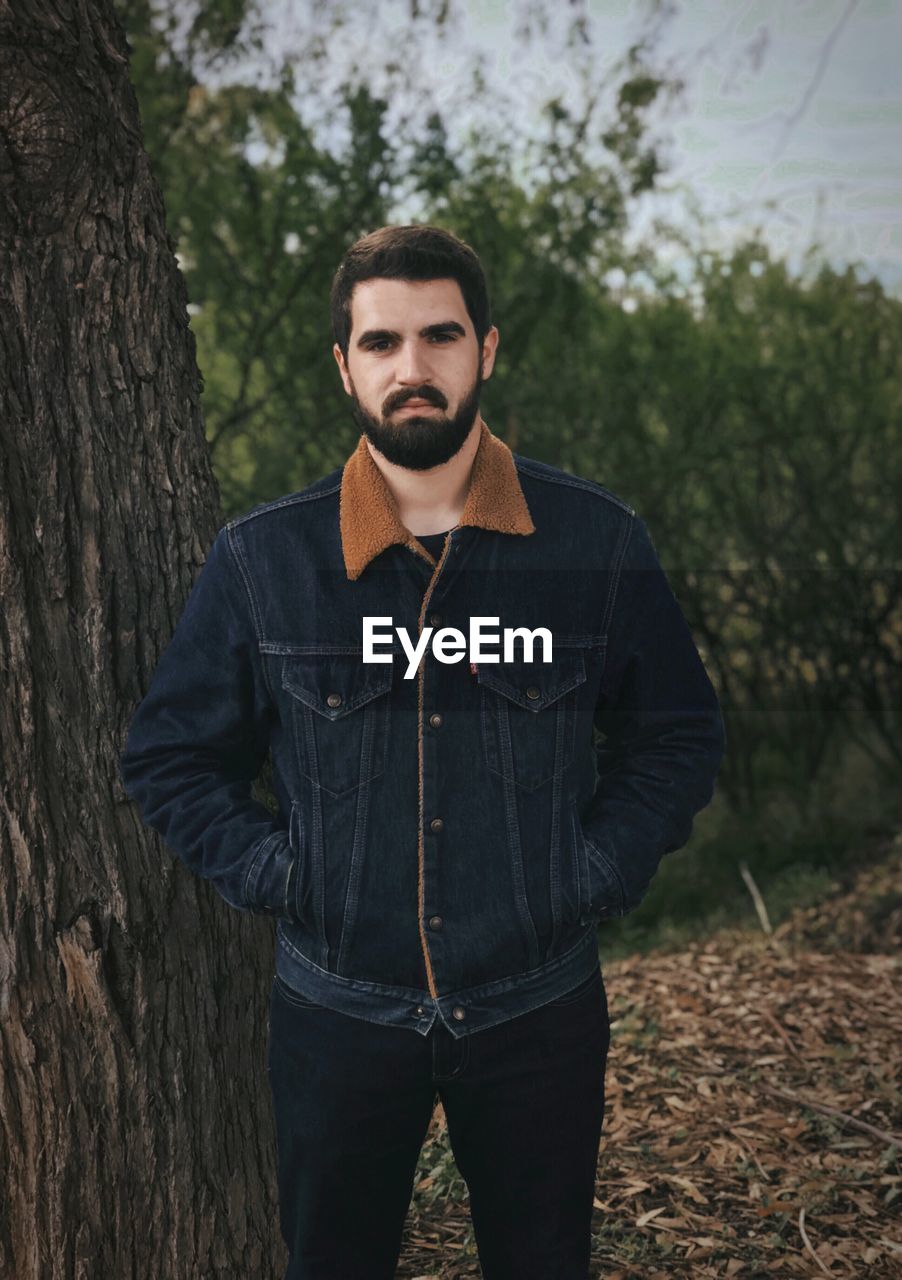 Portrait of handsome young man standing by tree trunk in forest