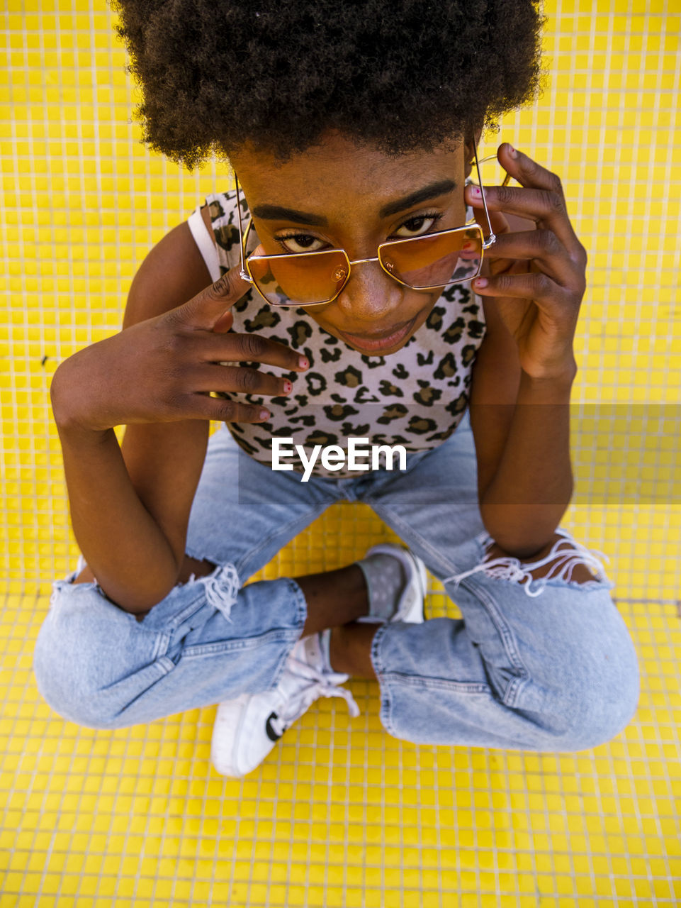 From above young african american female in trendy ripped jeans and stylish sunglasses sitting on yellow floor