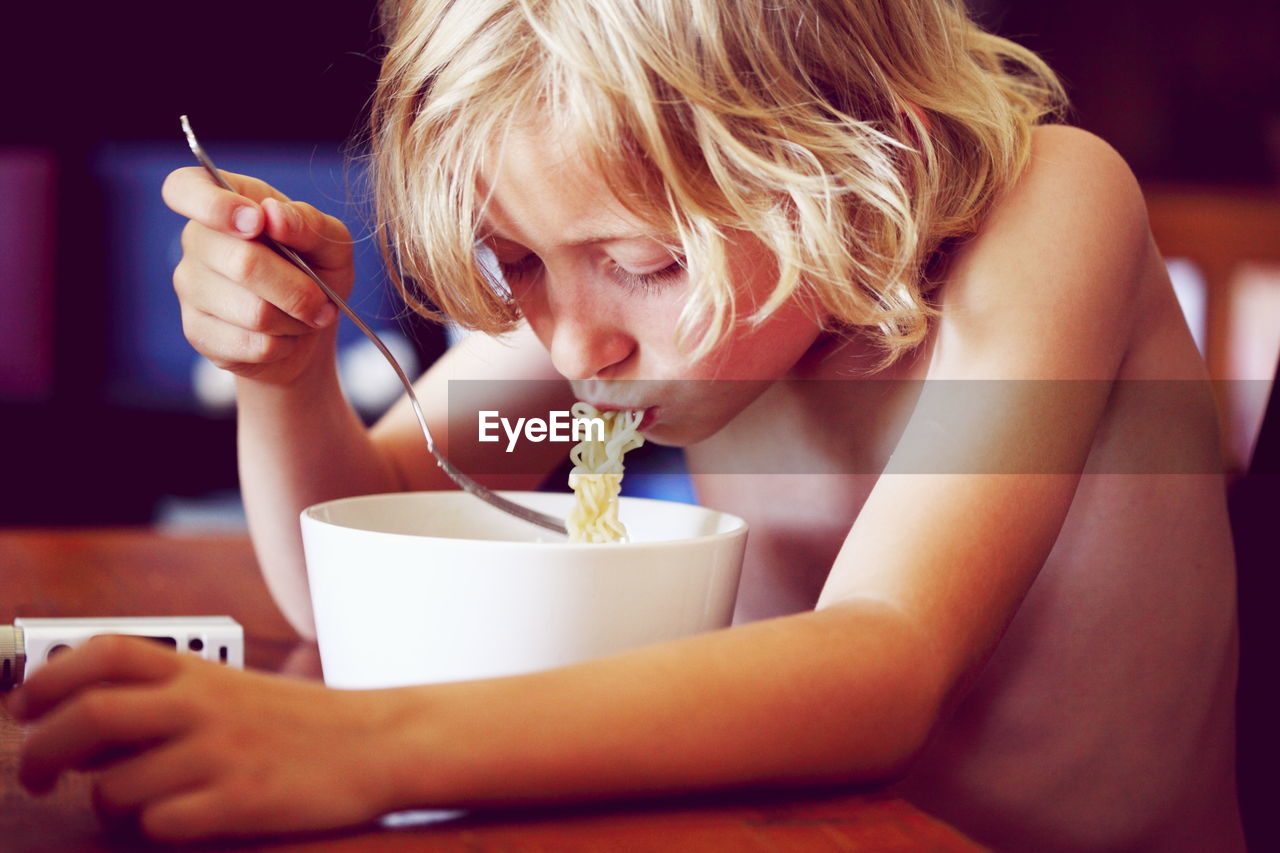 Shirtless blond boy eating noodles on table at home