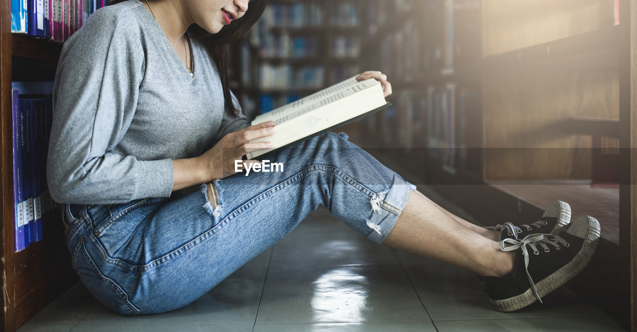 Side view of woman reading book while sitting on tiled floor in library