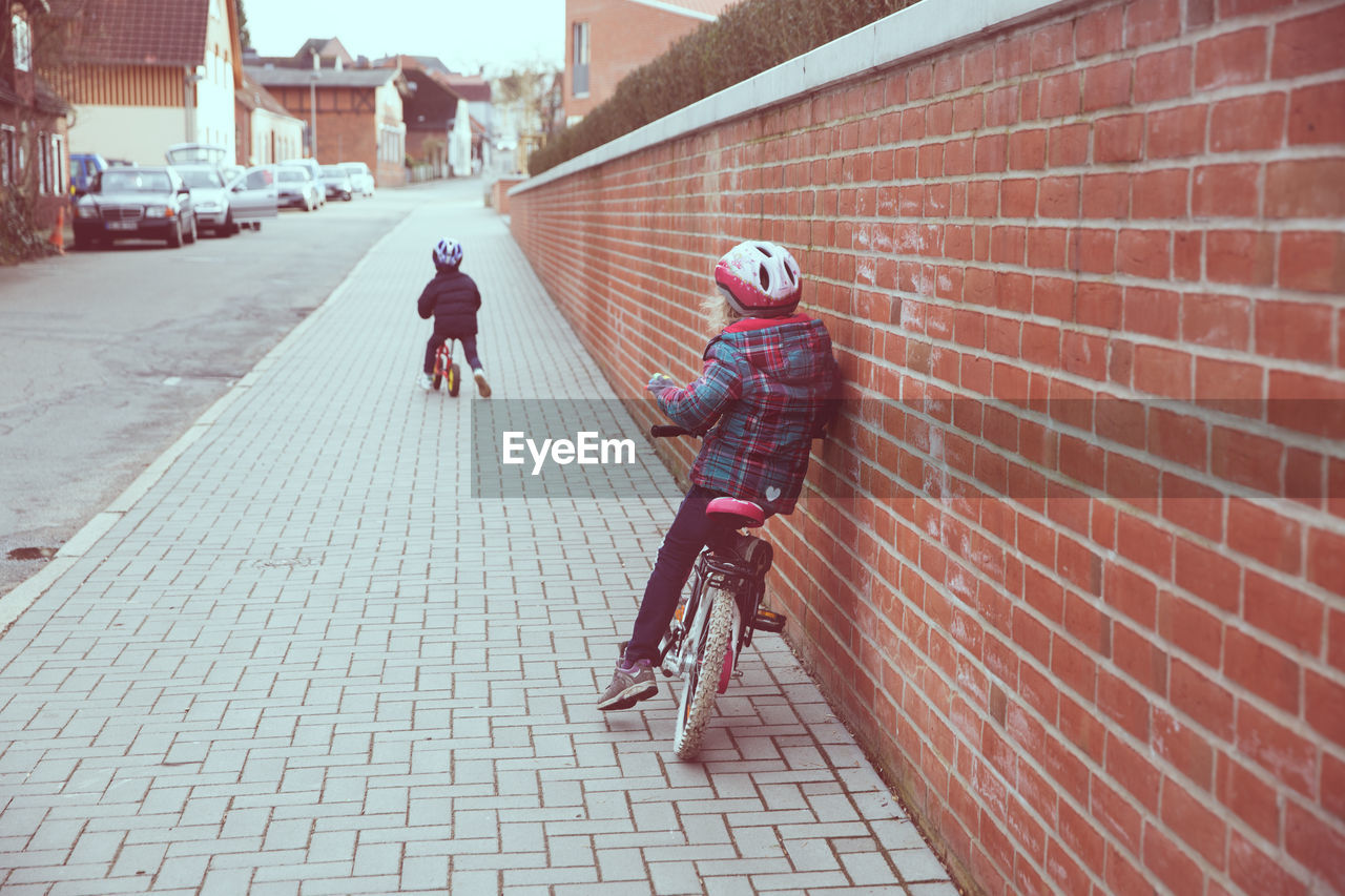 Children riding bicycle on sidewalk by brick wall