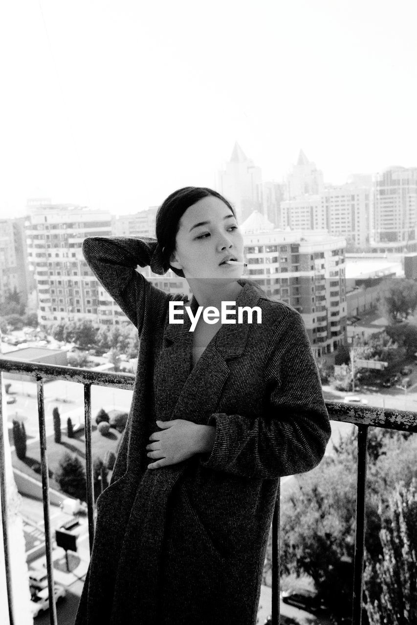 Young woman smoking while standing by railing in balcony against sky